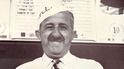 A black and white photo features a man sitting at a desk. He is wearing a hat and has a slight smile on his face. The setting appears to be an office or library, as suggested by the books visible in the background.
